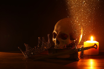 Human skull, old book, sword, crown and burning candle over old wooden table and dark background.