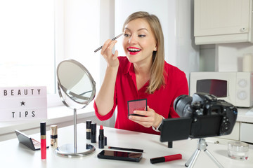 Stylish girl blogger in red blouse drawing on her eyebrows, looking at mirror, while recording make...