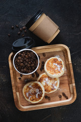 Takeaway coffee cups with coffee beans and curd fritters or syrniki on a wooden serving tray, top view on a dark brown stone background