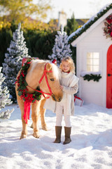 Nice blonde curly child caresses adorable pony with festive wreath near the small wooden house and snow-covered trees. New Year and Christmas time