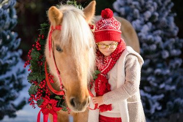 Close-up little boy with glasses and adorable pony with festive wreath near the small wooden house and snow-covered trees. New Year and Christmas time