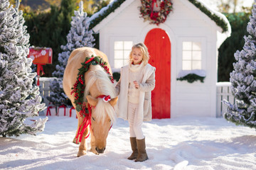 Nice blonde curly child and adorable pony with festive wreath near the small wooden house and...