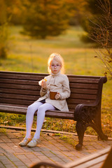 Curly schoolgirl in the glasses and light coat sitting on the branch in the autumn park and eating tasty cake on the sunset
