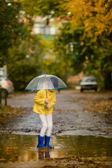 Child girl in yellow jacket and blue rubber boots with umbrella in puddle on an autumn walk