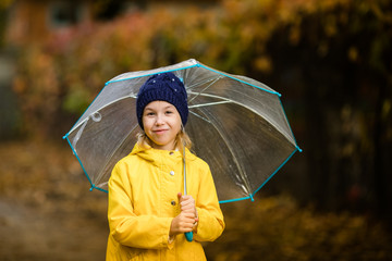 Child girl in yellow jacket and dark blue hat with transparent umbrella in the autumn park