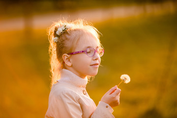 Curly schoolgirl in the glasses and light coat playing with dandelion in the autumn park on the sunset