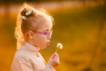 Curly schoolgirl in the glasses and light coat blowing at dandelion in the autumn park on the sunset