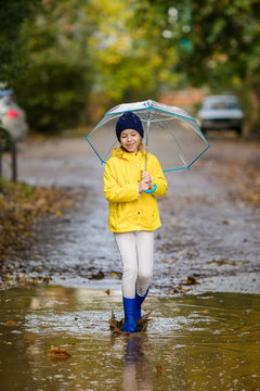 Child girl in yellow jacket and blue rubber boots with umbrella in puddle on an autumn walk