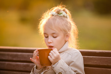 Nice curly child in the light coat with a big cup of hot cocoa in the autumn park