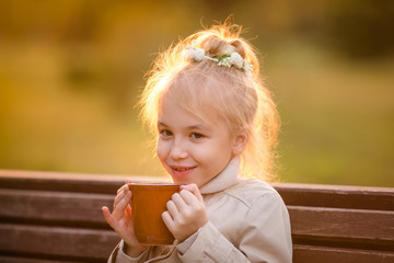 Nice curly smiling child in the light coat with a big cup of hot cocoa in the autumn park