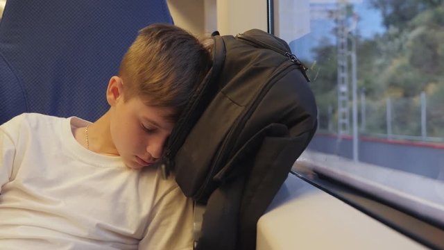 The boy is traveling by train sleeping near the window on his backpack. Sleep after a hard day traveler