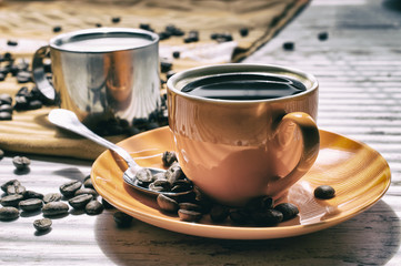 Coffee in an orange cup color. Milk in a metal cup.  Coffee in the side light. Spilled coffee beans. Still life in vintage style or in rustical style. Coffee on a pale oak table.