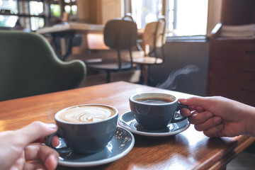 Closeup image of two people's hands holding coffee and hot chocolate cups on wooden table in cafe