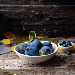 autumn background with ripe violet blue plums in white bowl on old grey wooden table, side view