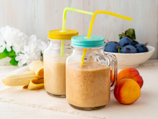 banana peach nectarine smoothie in two jars, fresh fruit milkshakes on white rustic wooden table, side view