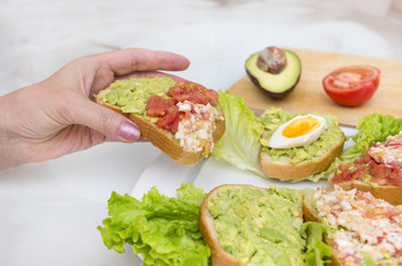hand holding a sandwich with avocado, tomato and egg, snack with avocado on lettuce leaves on a plate on the table