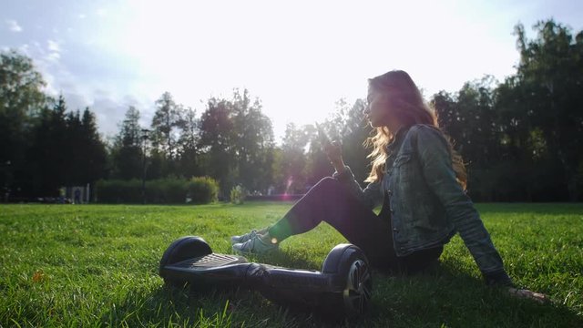 Cute young girl sitting on the grass, holding the phone in his hands. The gyroscope is next to her feet.