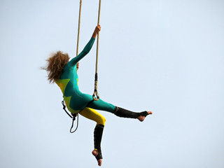 Girl aerial acrobat sitting on trapeze on sky background during a street circus performance. Woman...