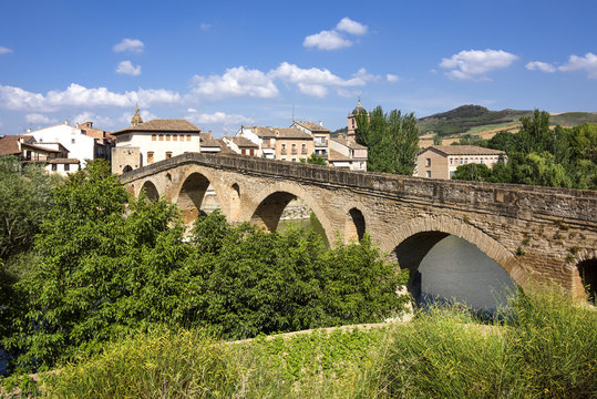 Fototapeta Spain, Puente La Reina, Gares: Panoramic view of famous romanesque bridge over river Arga with skyline of Spanish small town literally named 'Bridge of the Queen', green riverside and blue sky.