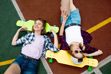 Two pretty smiling blond girls wearing checkered shirts, caps and denim shorts are lying on the bright longboards on the sportsfield with their eyes closed. Sport and cool style.