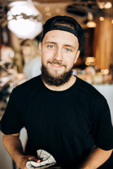 A handsome smiling youthful stylish man with beard,dressed in casual outfit,stands and looks at the camera in a cozy coffee shop.