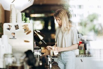A young pretty thin blonde with long hair,dressed in casual outfit,is cooking coffee in a modern coffee shop. Process of making coffee is shown.