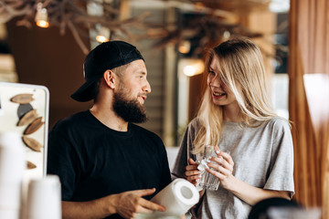 Two young smiling people,a blonde girl and man with beard.dressed in casual outfit, stand next to each other and look at each other  in a cozy coffee shop.