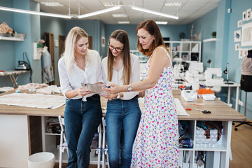 Three smart-looking pretty women wearing white shirts are looking at the sewing magazine. Fashion, tailor's workshop.