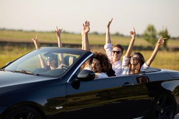 Beautiful young girls and guys in sunglasses smile and ride in a black cabriolet on the road holding their hands up on a sunny day.