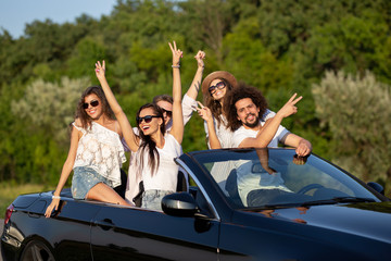 Lucky stylish young dark-haired girls and guys in sunglasses smile in a black cabriolet on the road...