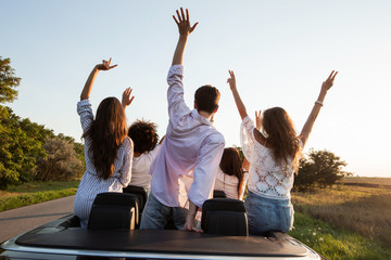 Back view. Young guys are sitting and holding hands up in a black cabriolet on the country road on...