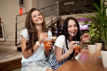 Two pretty youthful smiling girls,dressed in casual outfit,sit next to each other and look at the camera in a cozy coffee shop.