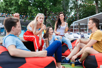 Obraz premium A company of good-looking friends laughing, drinking yellow cocktails and socialising while sitting on the bench and on the lawn in the nice summer cafe. Cheers. Entertainment, having good time