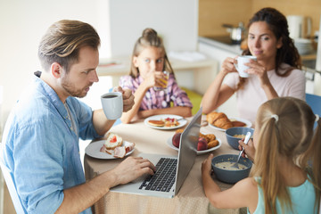 Young man searching for something in the net during breakfast with his wife and little daughters