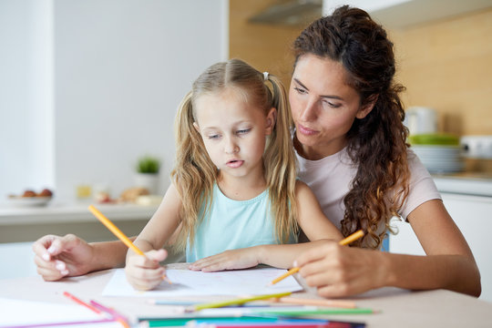 Young mother helping her little daughter with drawing picture with crayons at home