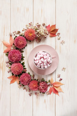 Hot drink with pink marshmallow in  pink cup surrounded by  floral pattern of pink flowers and leaves on white wooden background. Top view, close-up, place for text.