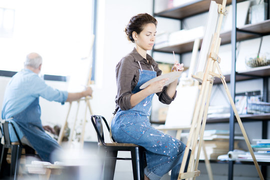 Young woman mixing colors on palette while sitting in front of easel with painting man on background