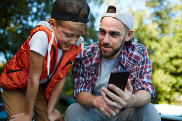 Young man with smartphone showing his son curious video about fishing or some other lesiure pastime