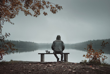 A man is sitting on a bench and looking at the lake. Back view. Also a coffee maker and a mug on...