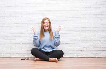 Young adult woman sitting on the floor in autumn over white brick wall celebrating mad and crazy for success with arms raised and closed eyes screaming excited. Winner concept