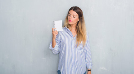 Young adult woman over grey grunge wall holding blank notebook with a confident expression on smart face thinking serious