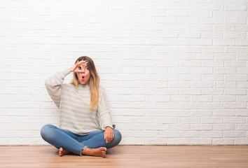 Fototapeta na wymiar Young adult woman sitting on the floor over white brick wall at home doing ok gesture shocked with surprised face, eye looking through fingers. Unbelieving expression.