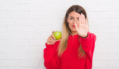 Young adult woman over white brick wall eating fresh green apple with open hand doing stop sign with serious and confident expression, defense gesture