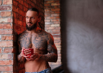 Fototapeta na wymiar Stylish shirtless bearded male with muscular and tattooed body holds cup of coffee while leaning on a wall and looks out the window.