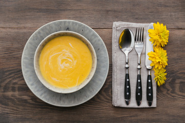Rustic table setting with pumpkin soup, linen napkin, cutlery, ceramic plates, yellow glasses and yellow flowers