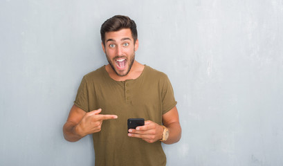 Handsome young man over grey grunge wall texting a message using smartphone very happy pointing with hand and finger