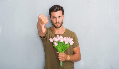 Handsome young man over grey grunge wall holding flowers bouquet with angry face, negative sign...