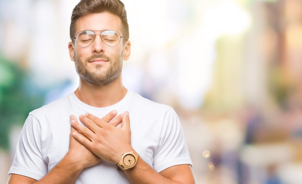 Young handsome man wearing glasses over isolated background smiling with hands on chest with closed eyes and grateful gesture on face. Health concept.