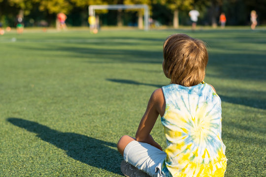 A Boy Is Sitting A Back On The Grass At The Edge Of A Stadium And Watching Football.