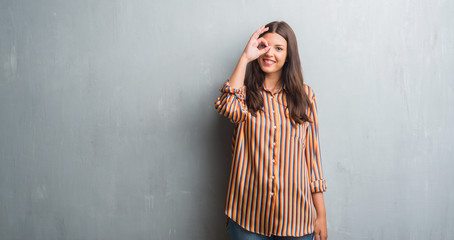 Fototapeta premium Young brunette woman over grunge grey wall doing ok gesture with hand smiling, eye looking through fingers with happy face.